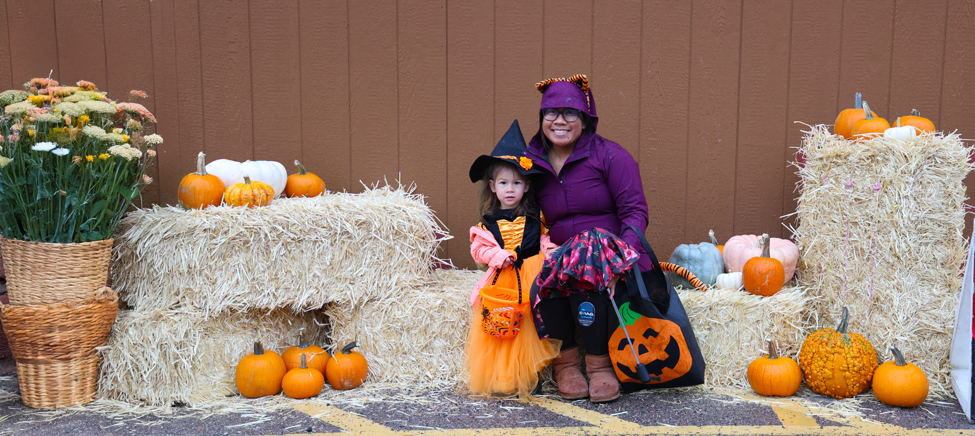 A mother poses with her daughter dressed as a witch on hay bales with pumpkins.