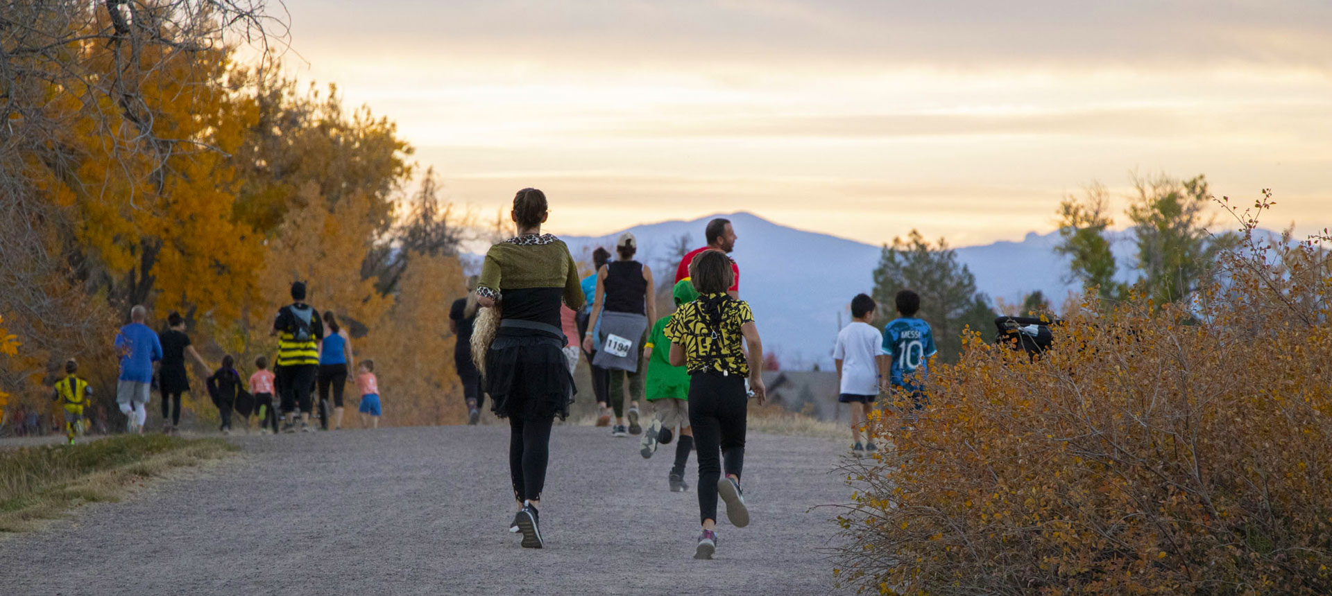 People of all ages walk and jog on a wide trail towards the mountains at sunset.