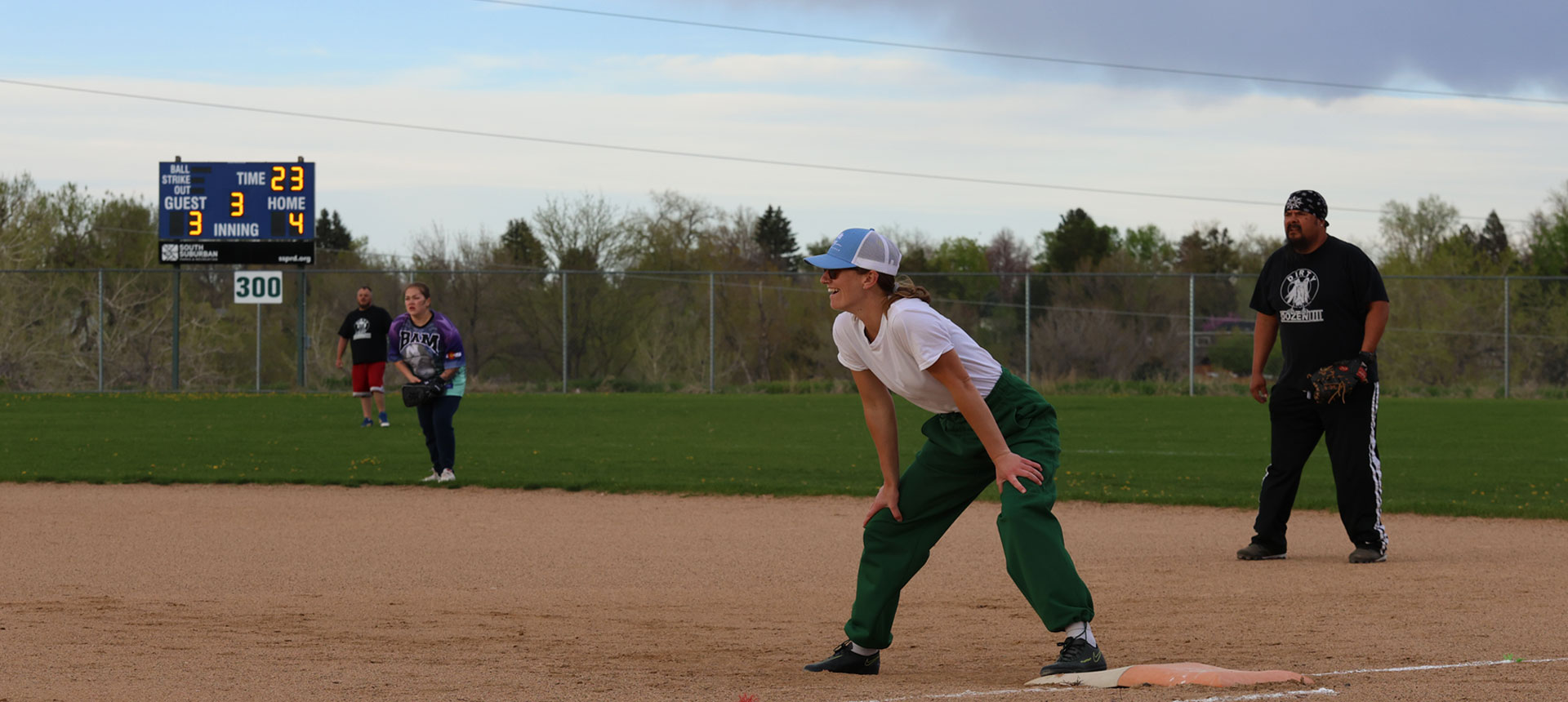 A smiling person stands on first base as 3 defensive softball players watch a play on a cloudy day.